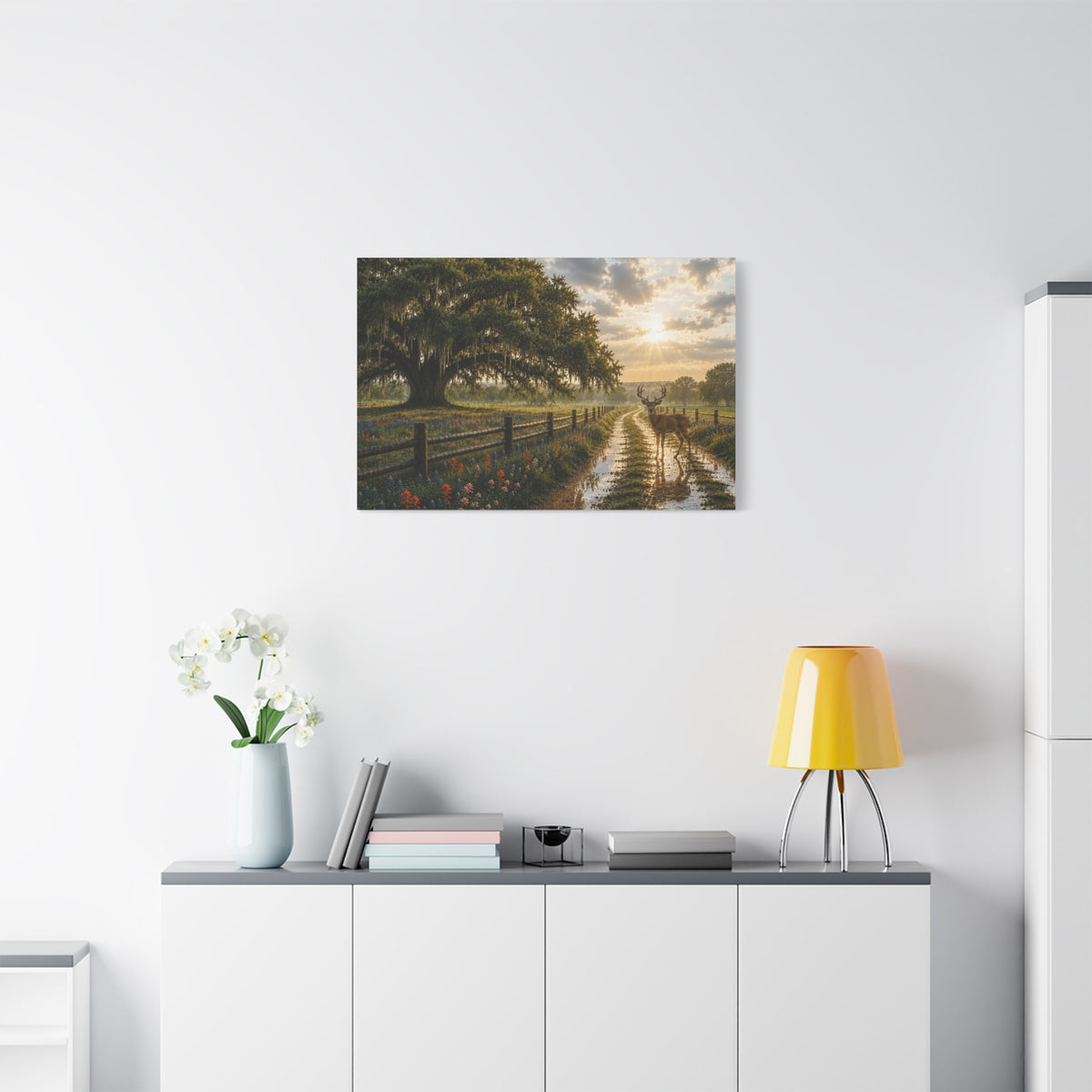 A landscape art print depicting a rustic country road with a large tree, a canal, and a sunset, displayed on a white wall above a modern white sideboard with a yellow lamp and white flowers.