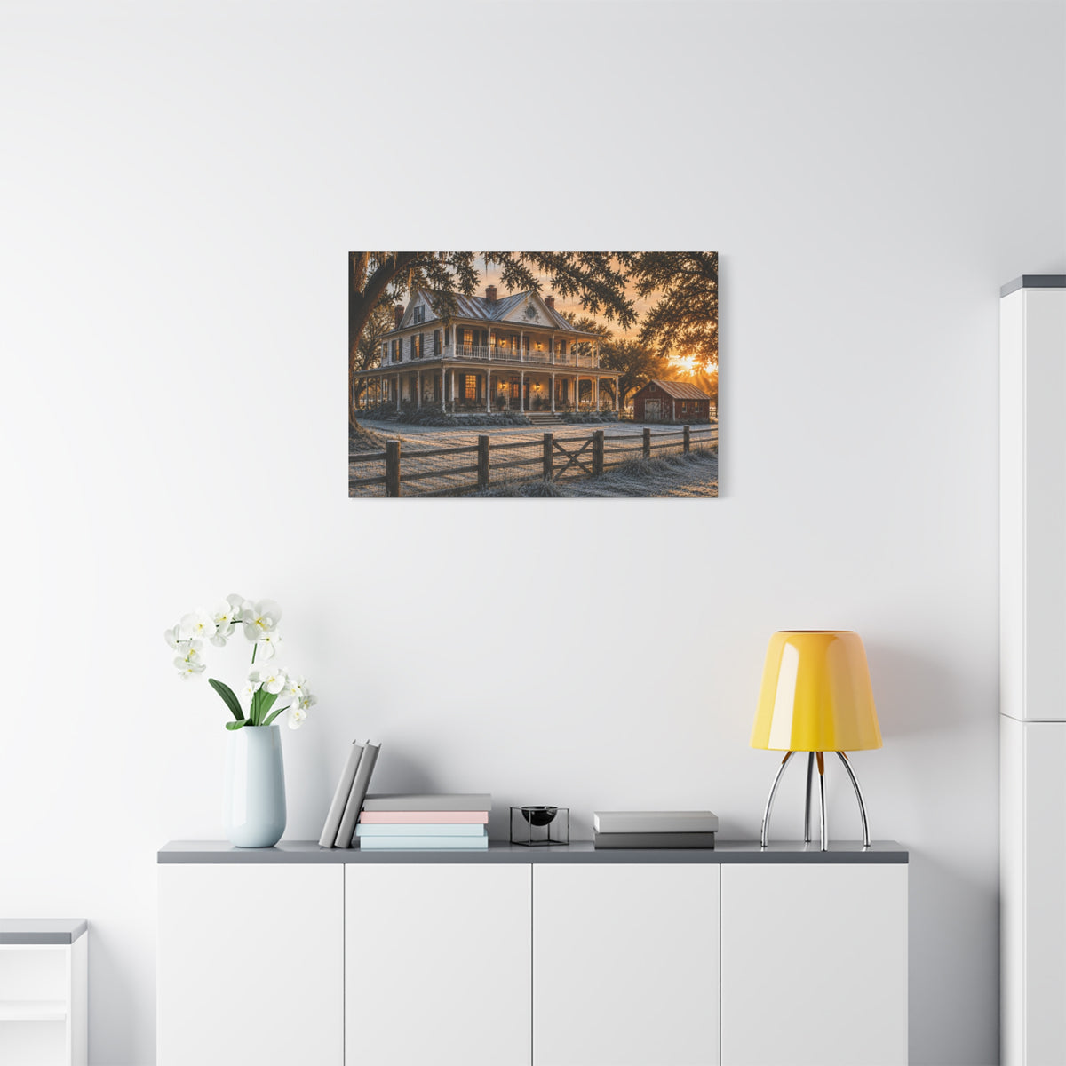 A First Frost canvas print of a frosted historic farmhouse at sunrise, displayed on a white wall above a console with a yellow lamp, books, and white flowers.