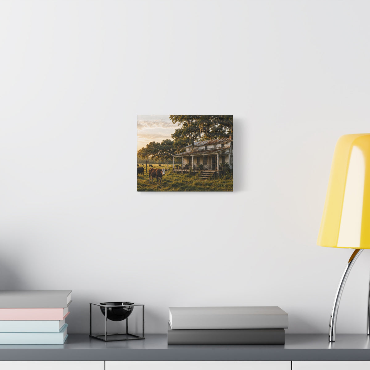 A square The Good Old Homestead canvas print showing a weathered two-story farmhouse with a front porch in a grassy pasture, with a cow in the foreground, cattle, moss-draped oak trees, and warm sunset light, displayed on a white wall above books and a yellow lamp.