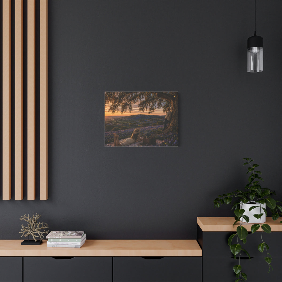 A framed wall art print depicting a sunset landscape with purple lavender fields and a tree, displayed on a white wall above a console table with a vase of white flowers, books, a small decor object, and a yellow lamp.