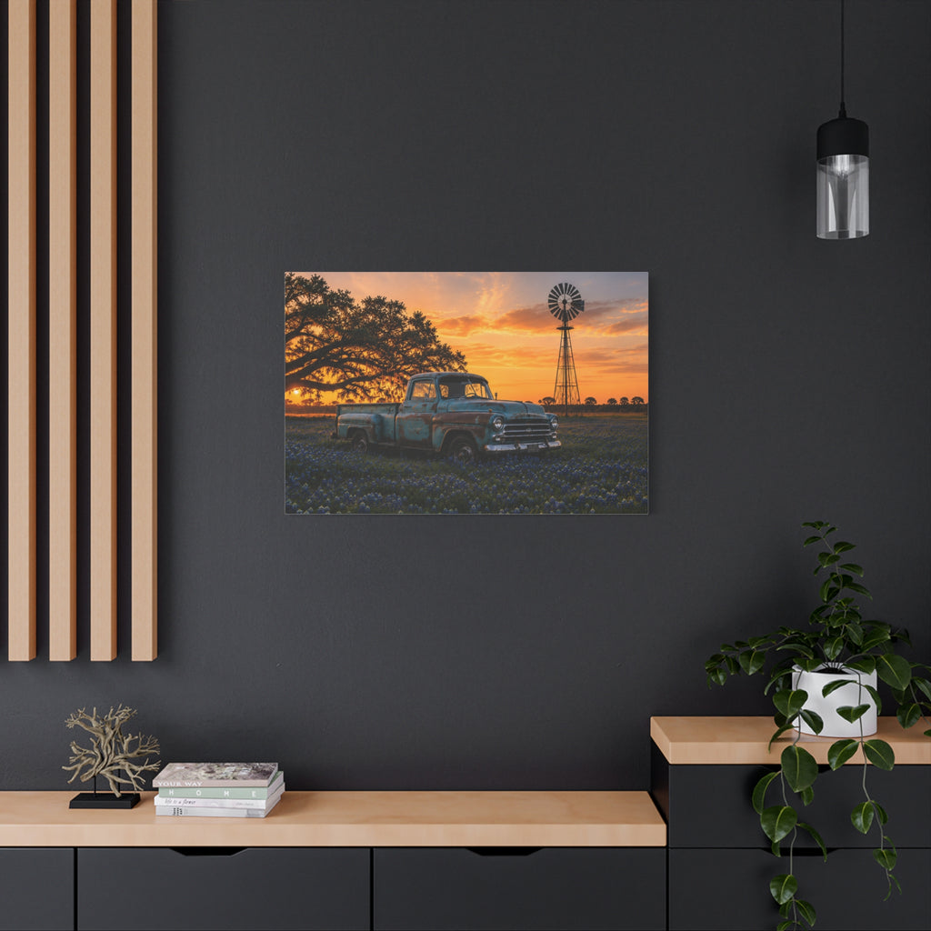 A The Windmill canvas print showing a weathered blue vintage pickup in a field of bluebonnets at sunset, with a windmill and large oak tree in the background, displayed on a white wall above a console with a yellow lamp, books, and white flowers.