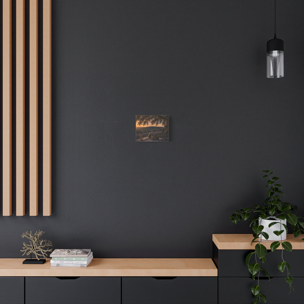A square wall art print depicting a scenic landscape with trees, hills, and a sunset, displayed on a white wall above a gray console table with books, a candle holder, and a yellow lamp.