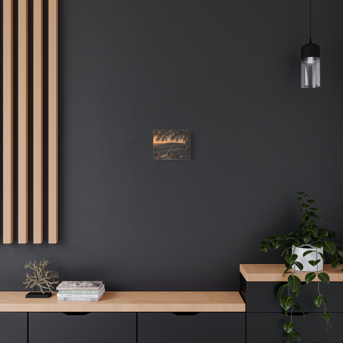 A square wall art print depicting a scenic landscape with trees, hills, and a sunset, displayed on a white wall above a gray console table with books, a candle holder, and a yellow lamp.