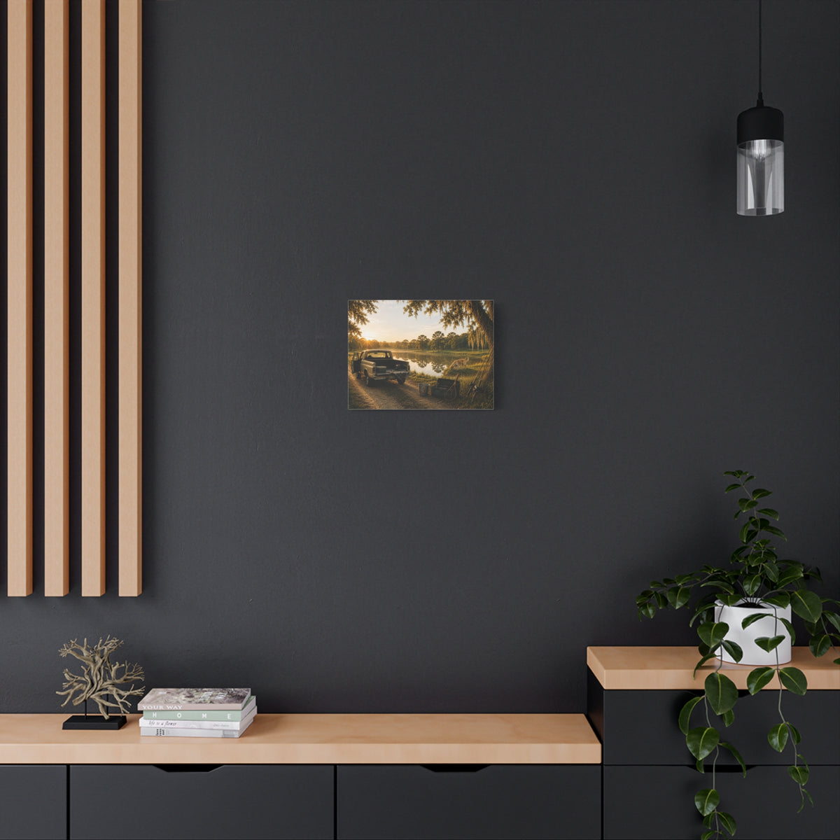 A The Old Fishing Hole canvas print showing a vintage pickup parked beside a quiet pond at sunset, with a dog, fishing rods, tackle, a cooler, and a moss-draped tree, displayed on a dark gray wall above a wooden console with a plant and hanging light.