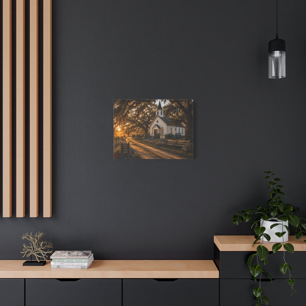 A square The Country Church canvas print showing a small white country church with a steeple beside a dirt path, framed by moss-draped oak trees, rustic benches, wildflowers, and warm sunset light, displayed on a dark charcoal wall above a light wood console with books and a plant.