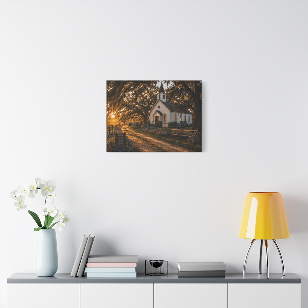 A The Country Church canvas print showing a small white country church with a steeple beside a dirt path, framed by moss-draped oak trees, rustic benches, wildflowers, and warm sunset light, displayed on a white wall above a console with a yellow lamp, books, and white flowers.