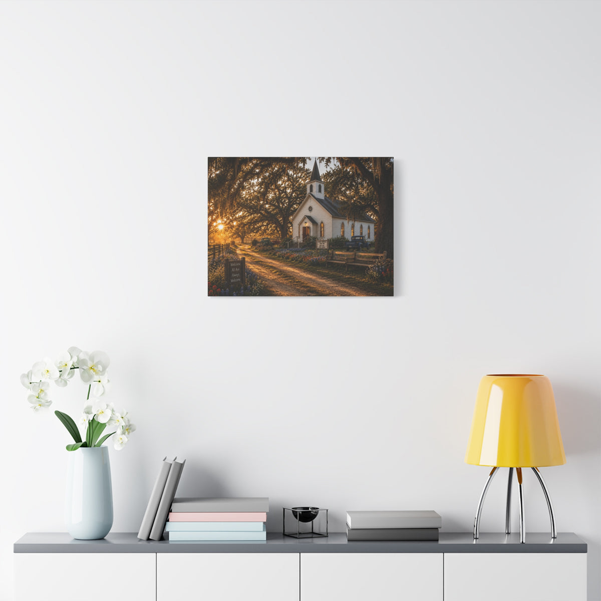 A The Country Church canvas print showing a small white country church with a steeple beside a dirt path, framed by moss-draped oak trees, rustic benches, wildflowers, and warm sunset light, displayed on a white wall above a console with a yellow lamp, books, and white flowers.