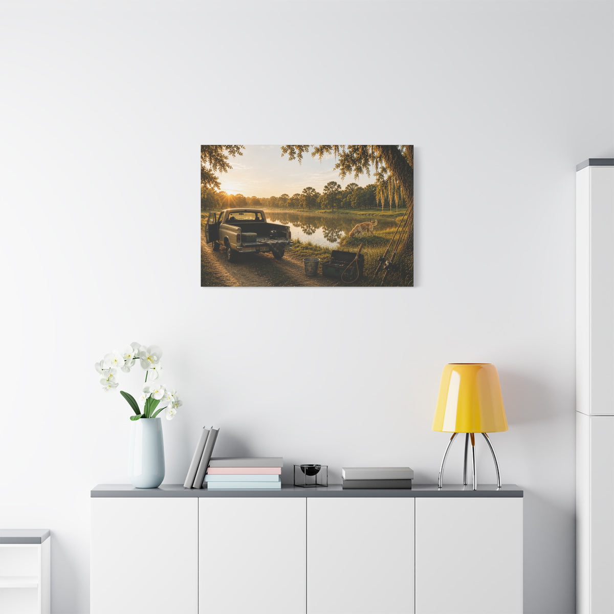 A The Old Fishing Hole canvas print showing a vintage pickup parked beside a quiet pond at sunset, with a dog, fishing rods, tackle, a cooler, and a moss-draped tree, displayed on a white wall above a console with a yellow lamp, books, and white flowers.