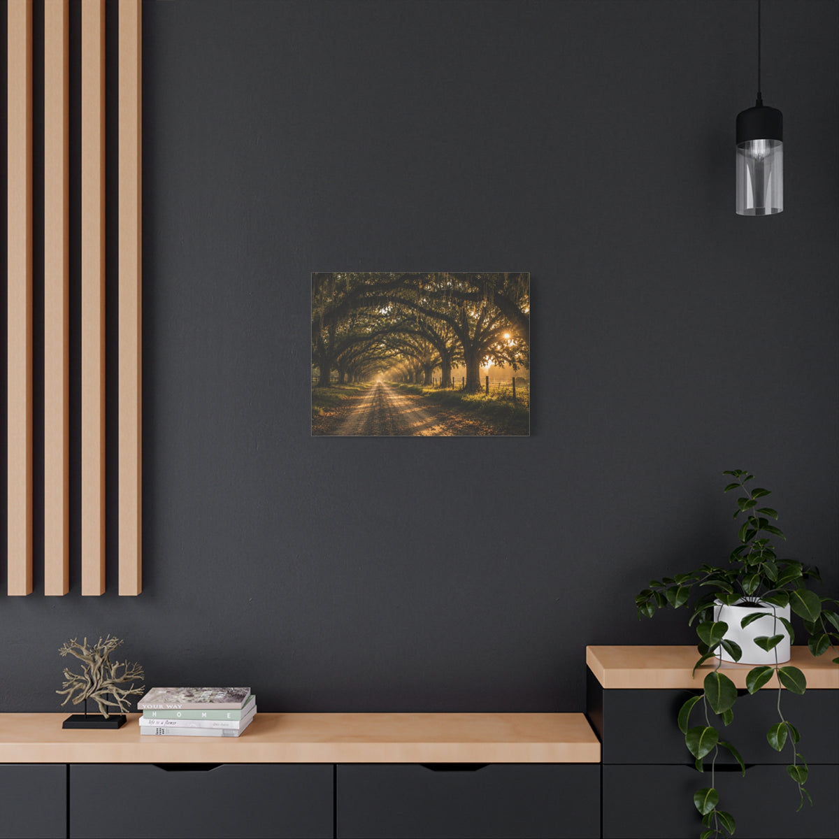 A square Sunday Morning canvas print showing a sunlit dirt road beneath a tunnel of moss-draped oak trees, with fences and warm morning light filtering through the branches, displayed on a dark charcoal wall above a light wood console with books and a plant.