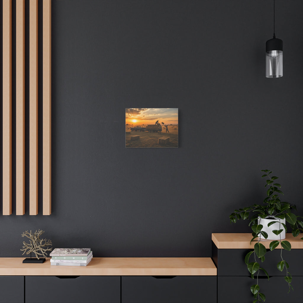 A Hay Season canvas print showing farmworkers loading rectangular hay bales onto a flatbed trailer beside a pickup truck in a golden field at sunset, displayed on a dark gray wall above a wooden console with a plant and hanging light.