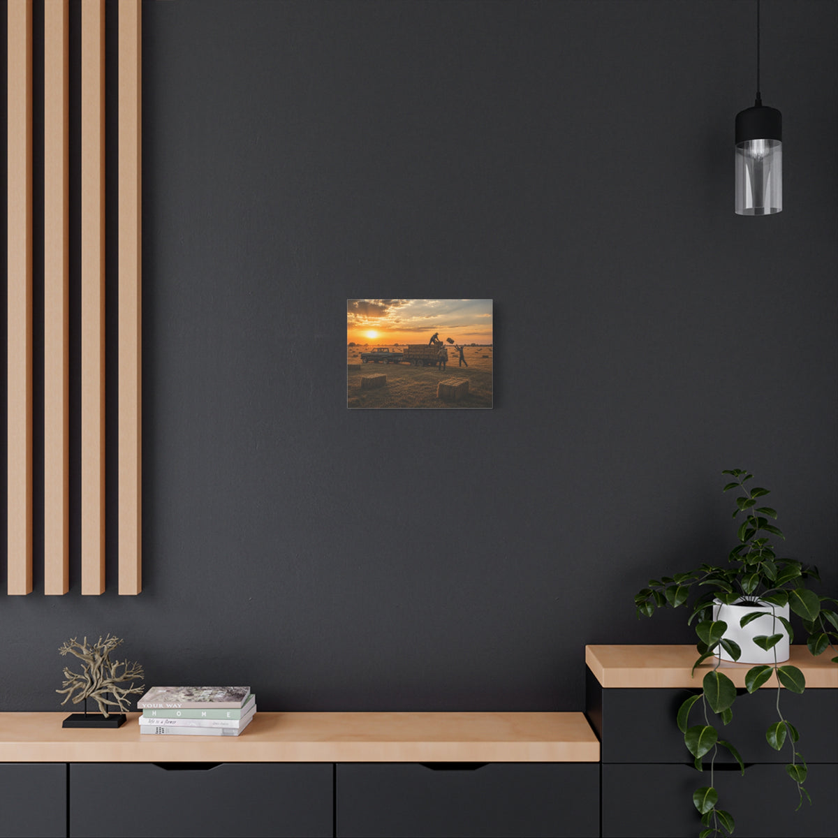 A Hay Season canvas print showing farmworkers loading rectangular hay bales onto a flatbed trailer beside a pickup truck in a golden field at sunset, displayed on a dark gray wall above a wooden console with a plant and hanging light.