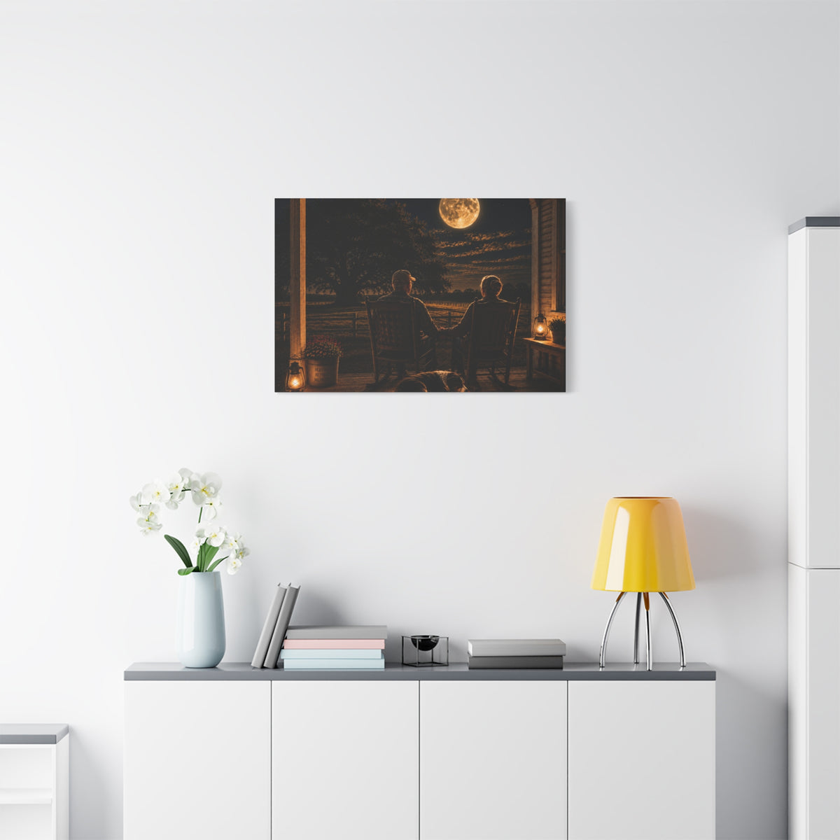 A Harvest Moon canvas print showing an older couple holding hands in rocking chairs on a farmhouse porch with a sleeping dog, glowing lanterns, and a full moon over the fields, displayed on a white wall above a console with a yellow lamp, books, and white flowers.