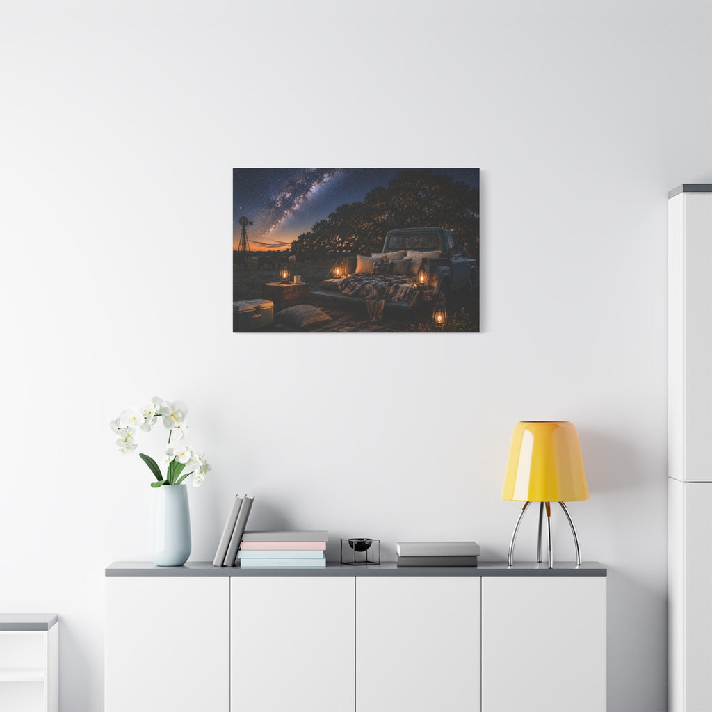 A Starry Night canvas print showing a cozy outdoor bed beside a vintage pickup in a country field, with lanterns, pillows, blankets, windmills, and the Milky Way overhead, displayed on a white wall above a console with a yellow lamp, books, and white flowers.