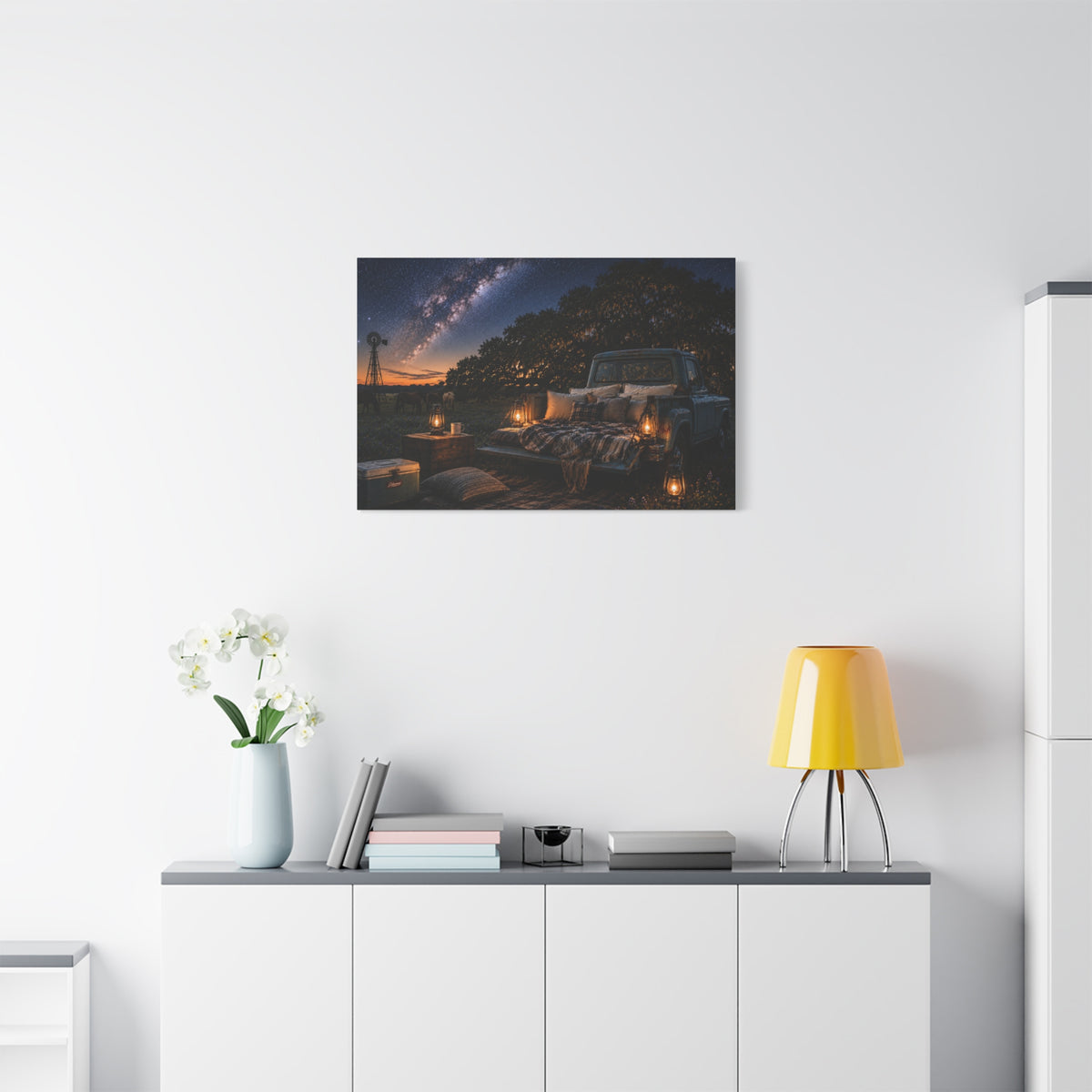 A Starry Night canvas print showing a cozy outdoor bed beside a vintage pickup in a country field, with lanterns, pillows, blankets, windmills, and the Milky Way overhead, displayed on a white wall above a console with a yellow lamp, books, and white flowers.