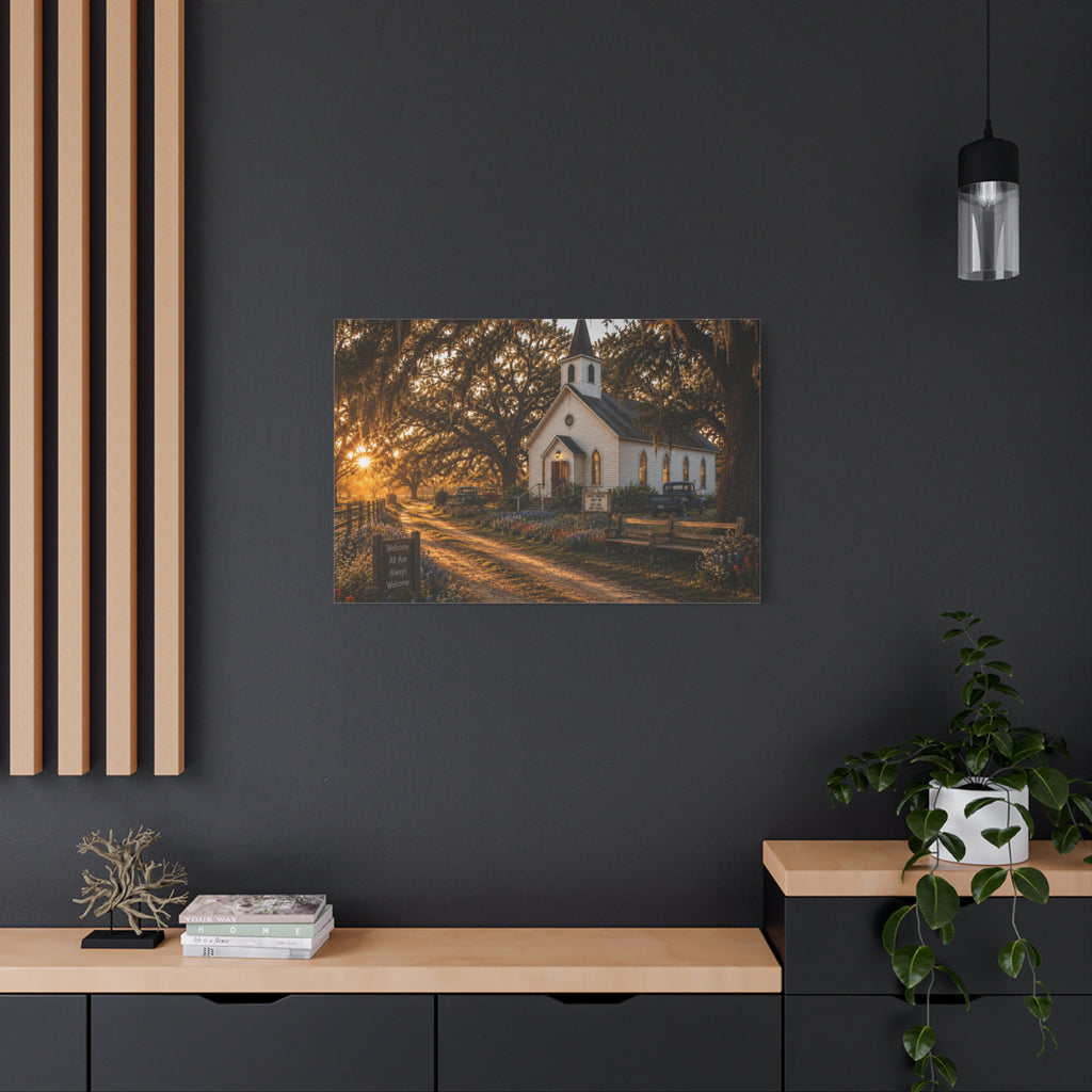 A rectangular The Country Church canvas print showing a small white country church with a steeple beside a dirt path, framed by moss-draped oak trees, rustic benches, wildflowers, and warm sunset light, displayed on a dark charcoal wall above a light wood console with books, a plant, and decor.