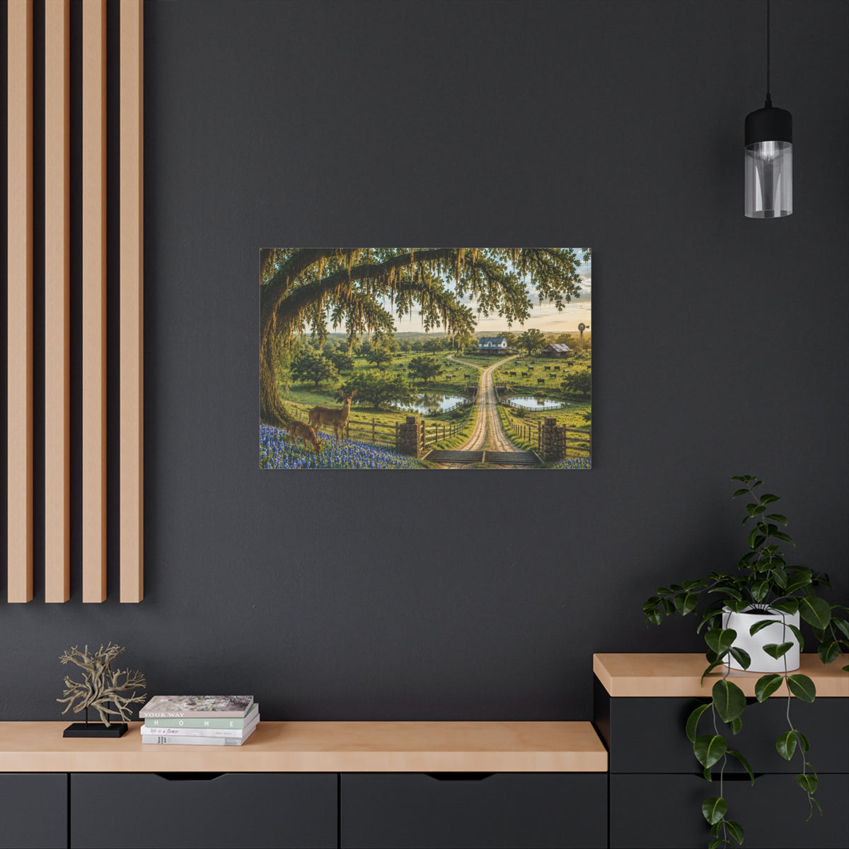 A framed landscape art print depicting a country road through a lush green valley under a large tree, displayed on a dark gray wall in a modern styled room with a wooden console, a potted plant, and a hanging light.