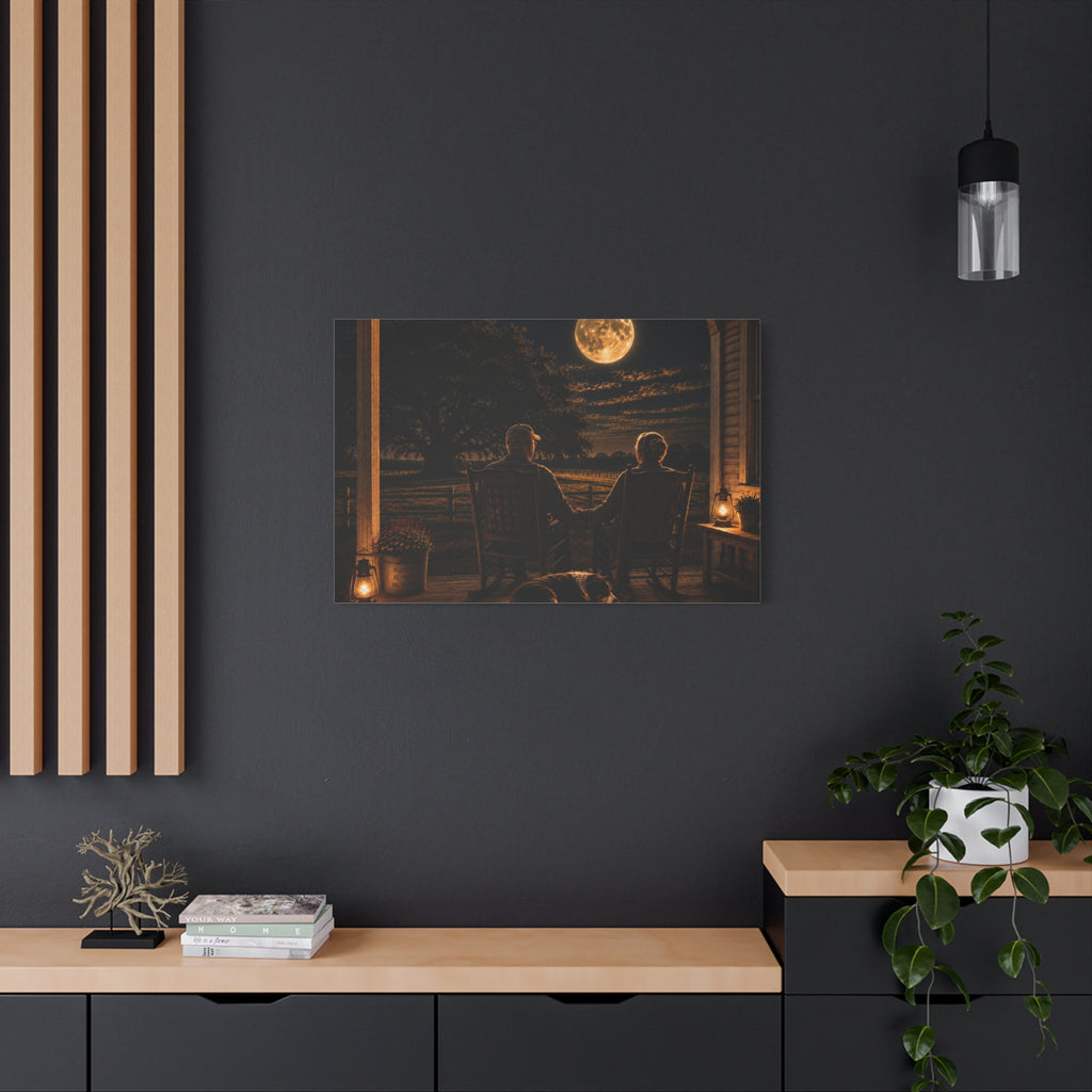 A rectangular Harvest Moon canvas print showing an older couple holding hands in rocking chairs on a farmhouse porch with a sleeping dog, glowing lanterns, and a full moon over the fields, displayed on a dark charcoal wall above a light wood console with books, a plant, and decor.