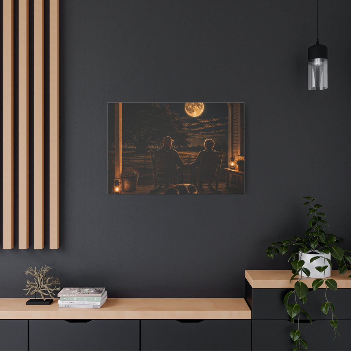 A rectangular Harvest Moon canvas print showing an older couple holding hands in rocking chairs on a farmhouse porch with a sleeping dog, glowing lanterns, and a full moon over the fields, displayed on a dark charcoal wall above a light wood console with books, a plant, and decor.