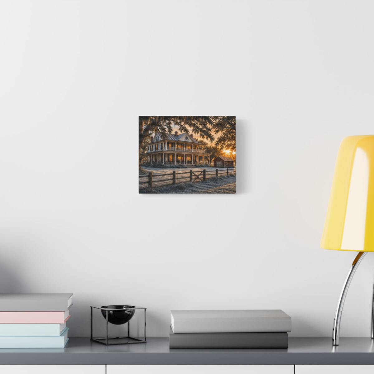 A square First Frost canvas print of a frosted historic farmhouse at sunrise, displayed on a white wall above a gray console with books, a black bowl, and a yellow lamp.