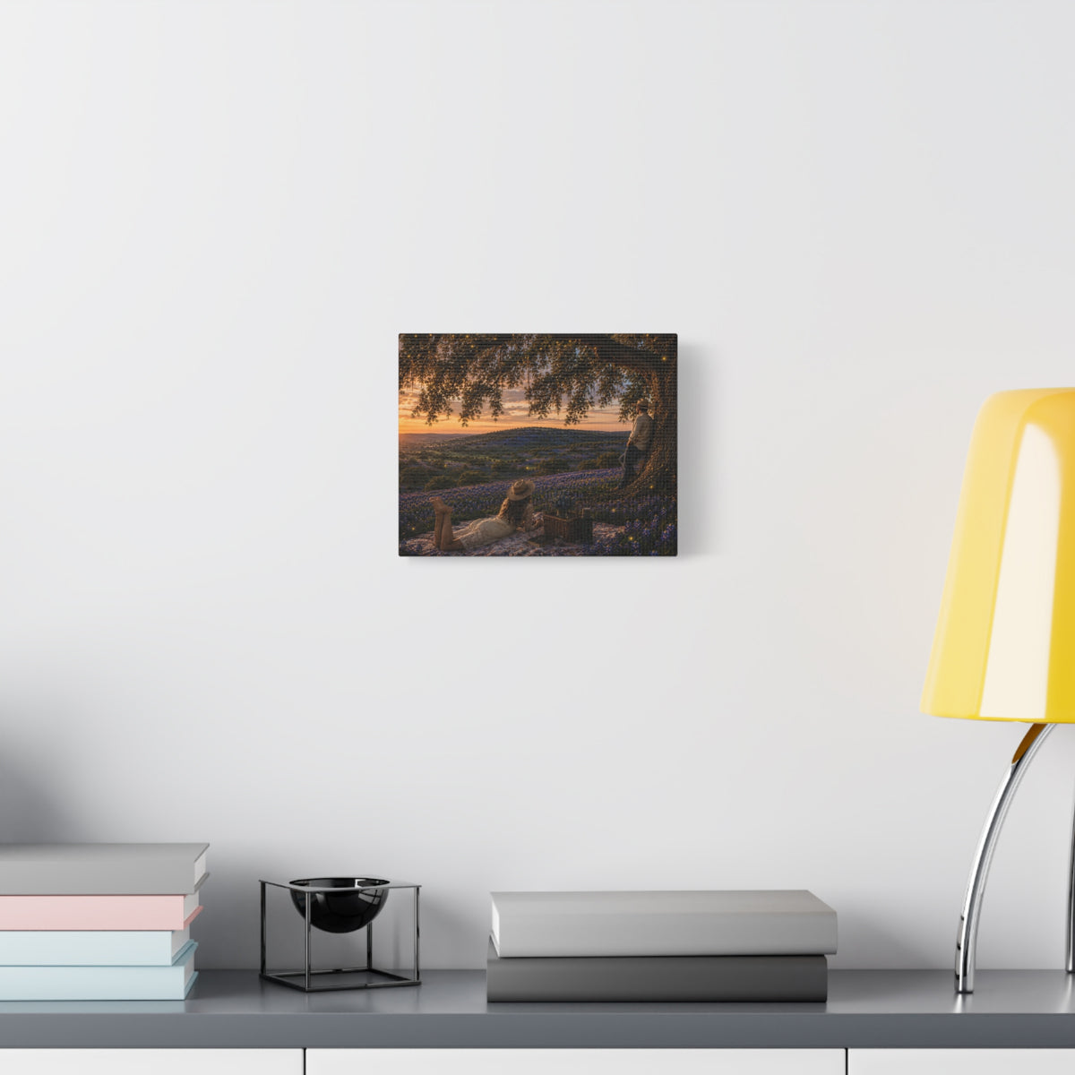 A square wall art print depicting a scenic landscape with trees, hills, and a sunset, displayed on a white wall above a gray console table with books, a candle holder, and a yellow lamp.