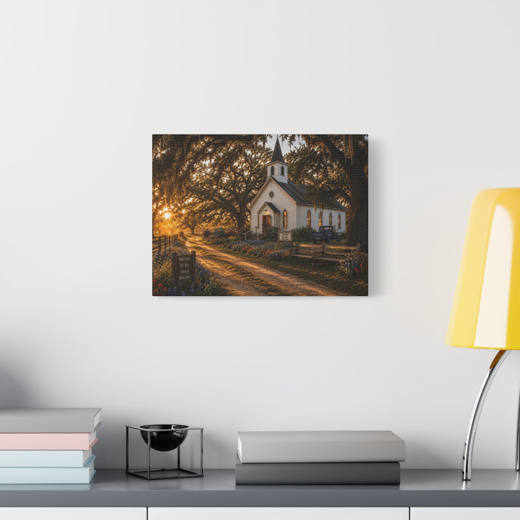 A rectangular The Country Church canvas print showing a small white country church with a steeple beside a dirt path, framed by moss-draped oak trees, rustic benches, wildflowers, and warm sunset light, displayed on a white wall above books, a black accent, and a yellow lamp.