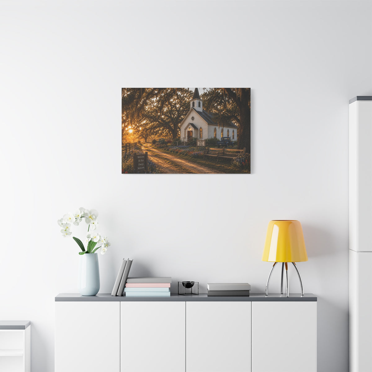 A The Country Church canvas print showing a small white country church with a steeple beside a dirt path, framed by moss-draped oak trees, rustic benches, wildflowers, and warm sunset light, displayed on a white wall above a console with a yellow lamp, books, and white flowers.