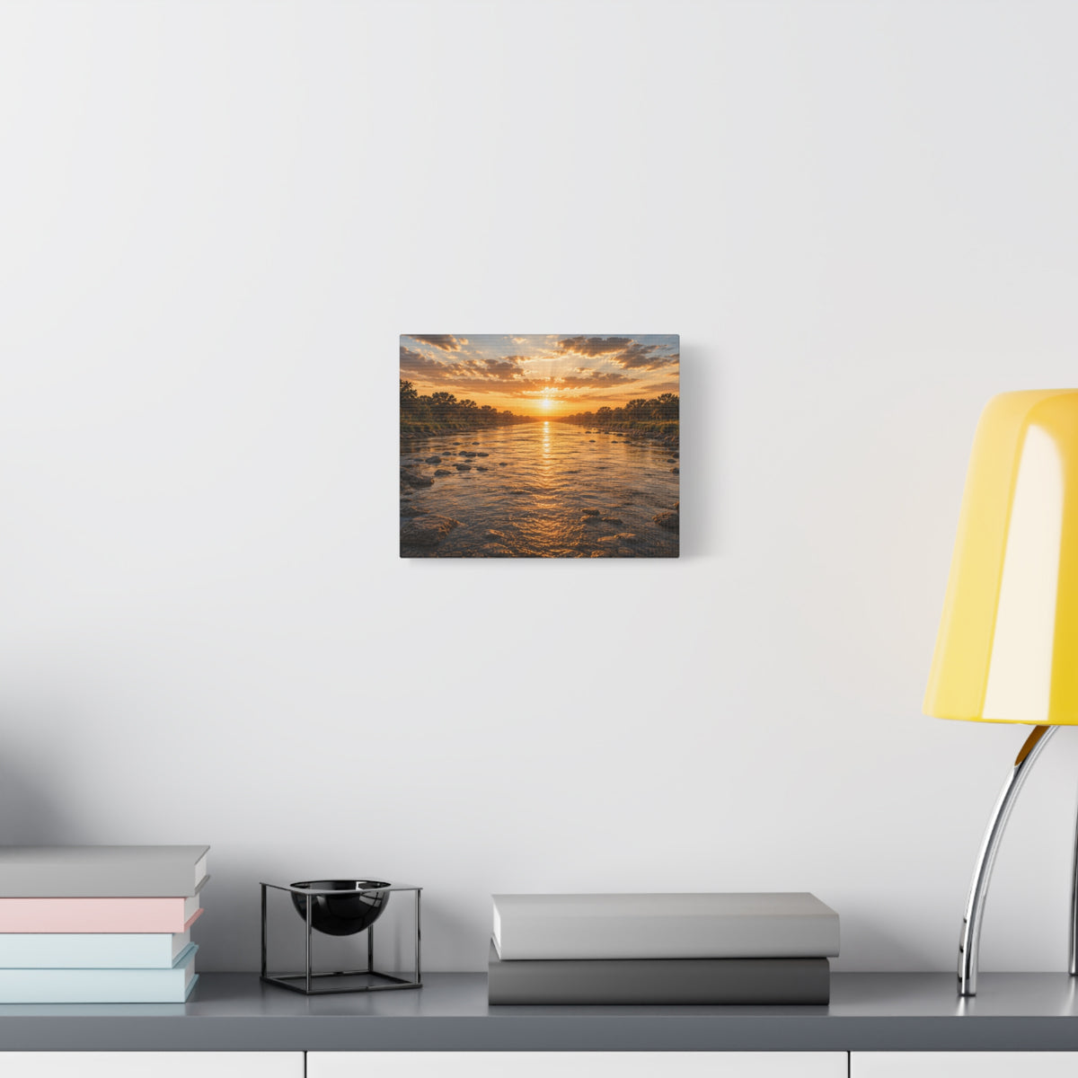 A The River canvas print showing a golden sunset over a shallow river, with rocks in the water, tree-lined banks, glowing reflections, and sunbeams breaking through the clouds, displayed on a dark wall above wooden shelves with a plant and decor.