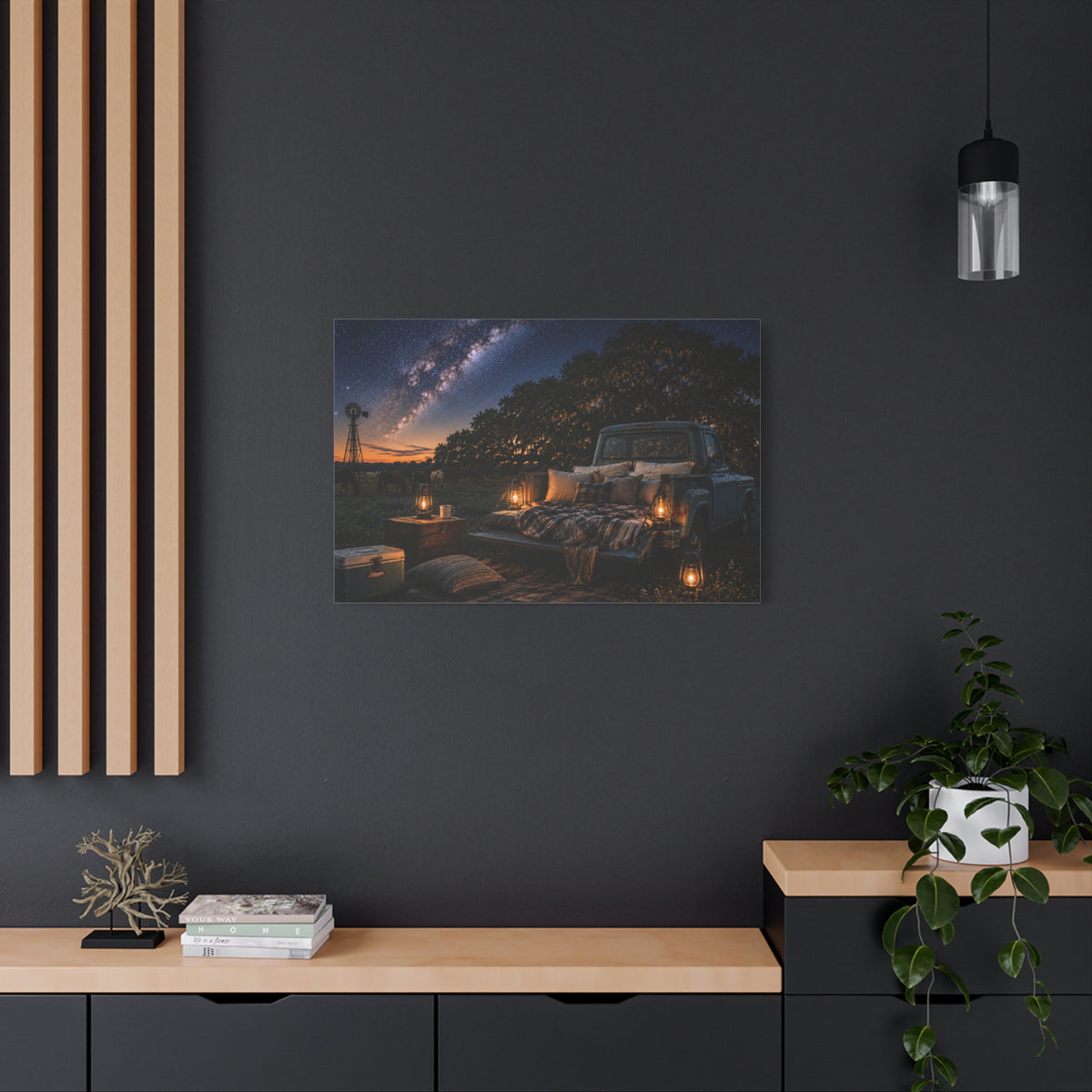 A rectangular Starry Night canvas print showing a cozy outdoor bed beside a vintage pickup in a country field, with lanterns, pillows, blankets, windmills, and the Milky Way overhead, displayed on a dark charcoal wall above a light wood console with books, a plant, and decor.