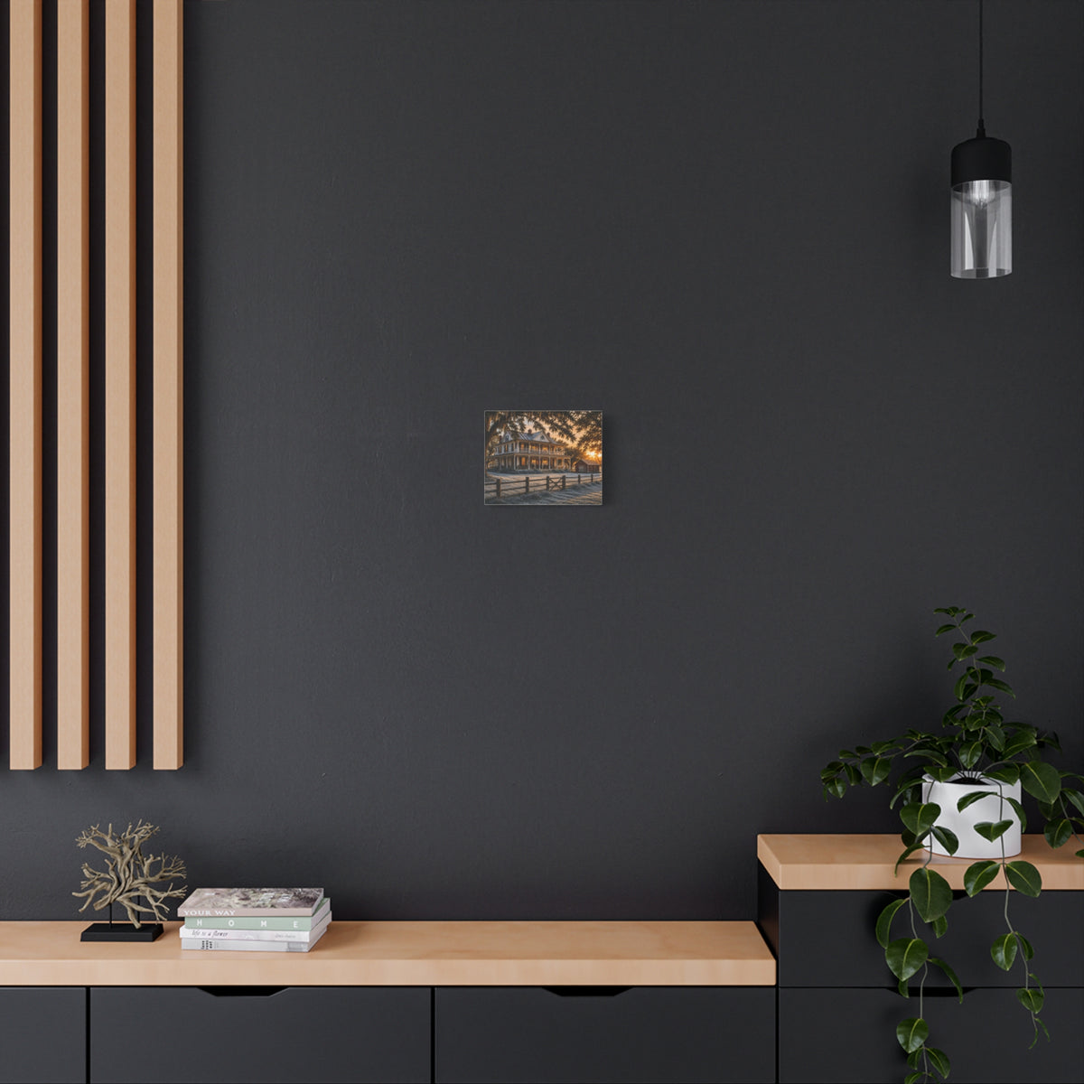A First Frost canvas print showing a historic farmhouse, frosted yard, fence, and warm sunrise light, displayed on a dark wall above a wooden shelf with a plant and decor.