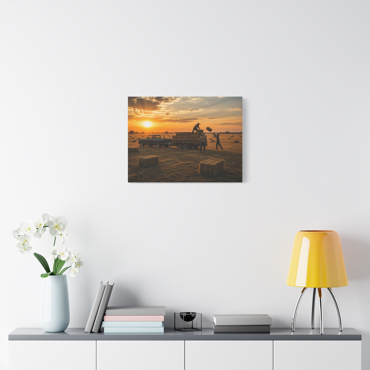 A Hay Season canvas print showing farmworkers loading rectangular hay bales onto a flatbed trailer beside a pickup truck in a golden field at sunset, displayed on a white wall above a console with a yellow lamp, books, and white flowers.