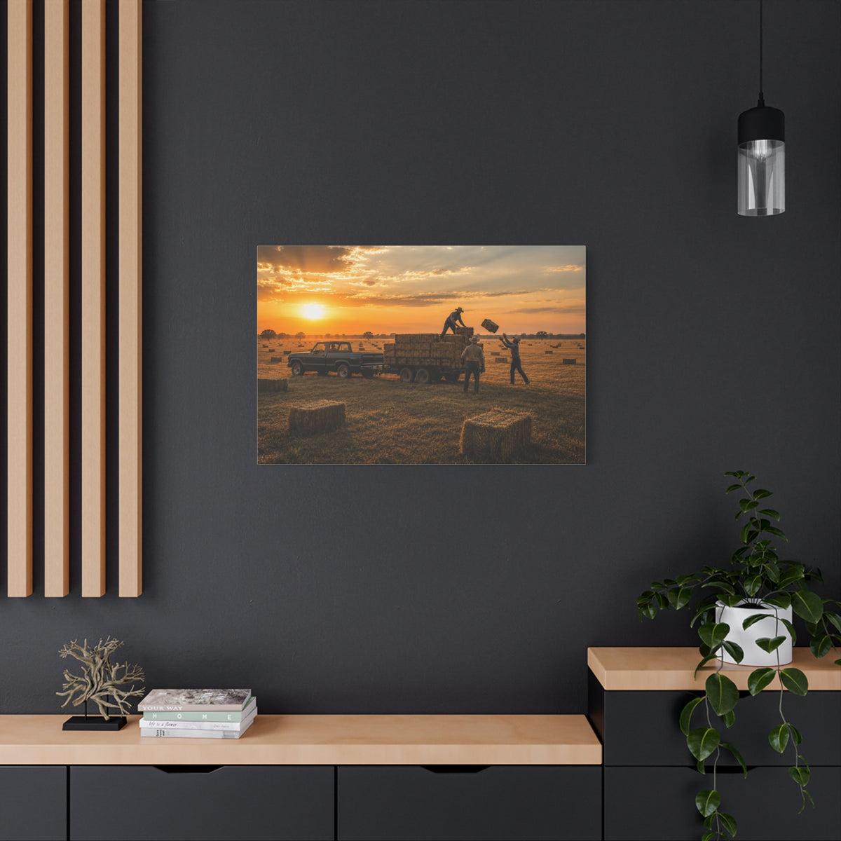 A rectangular Hay Season canvas print showing farmworkers loading rectangular hay bales onto a flatbed trailer beside a pickup truck in a golden field at sunset, displayed on a dark charcoal wall above a light wood console with books, a plant, and decor.