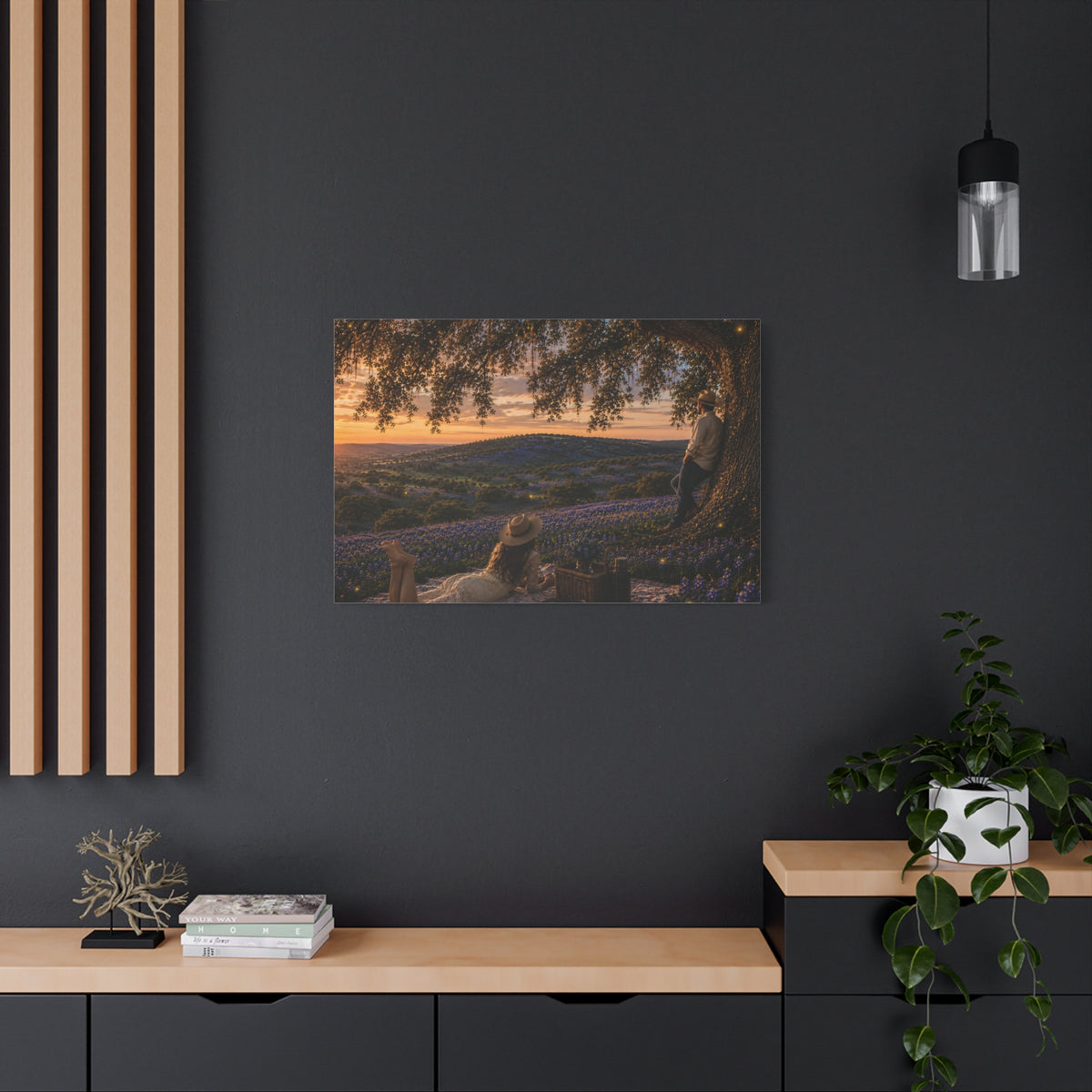 A framed landscape art print depicting a sunset over lavender fields under a tree, displayed on a dark gray wall in a modern room setting with a wooden console, potted plant, and hanging light.