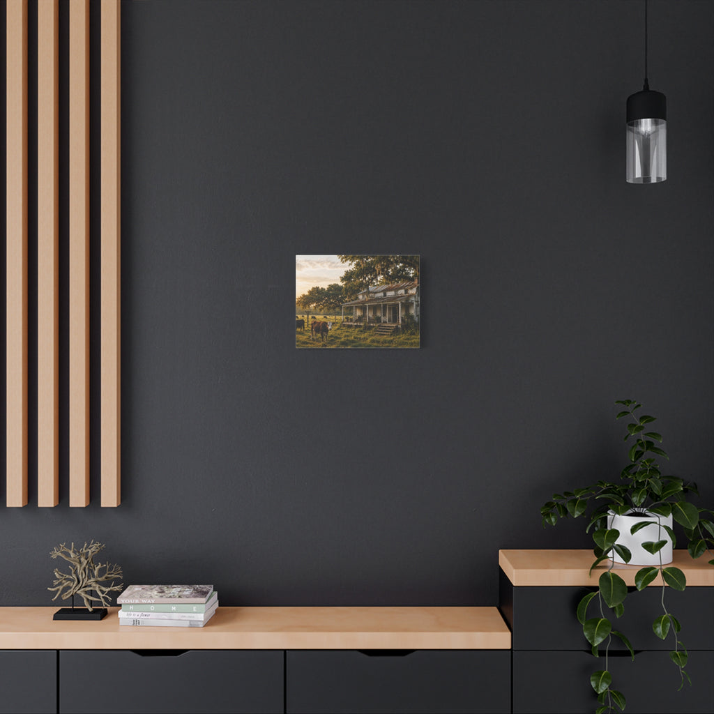 A The Good Old Homestead canvas print showing a weathered two-story farmhouse with a front porch in a grassy pasture, with a cow in the foreground, cattle, moss-draped oak trees, and warm sunset light, displayed on a dark gray wall above a wooden console with a plant and hanging light.
