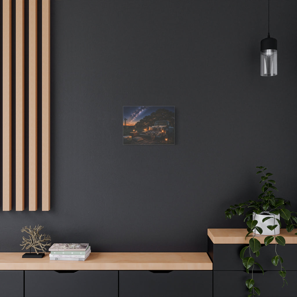 A Starry Night canvas print showing a cozy outdoor bed beside a vintage pickup in a country field, with lanterns, pillows, blankets, windmills, and the Milky Way overhead, displayed on a dark gray wall above a wooden console with a plant and hanging light.
