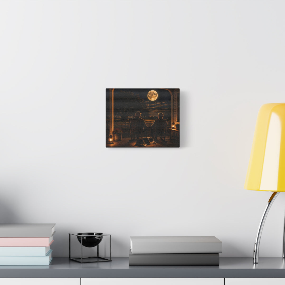 A square Harvest Moon canvas print showing an older couple holding hands in rocking chairs on a farmhouse porch with a sleeping dog, glowing lanterns, and a full moon over the fields, displayed on a white wall above books and a yellow lamp.
