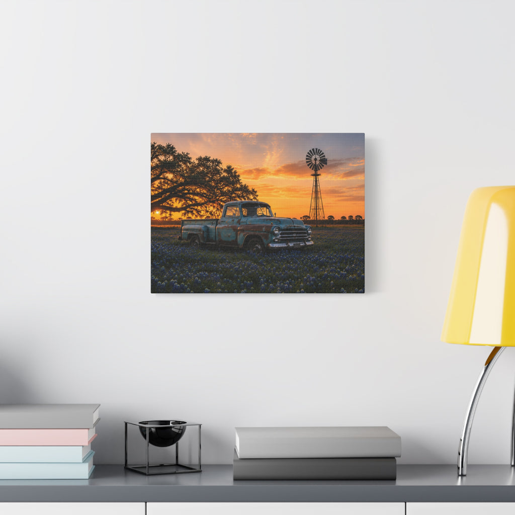 A square The Windmill canvas print showing a weathered blue vintage pickup in a field of bluebonnets at sunset, with a windmill and large oak tree in the background, displayed on a dark charcoal wall above a light wood console with books and a plant.