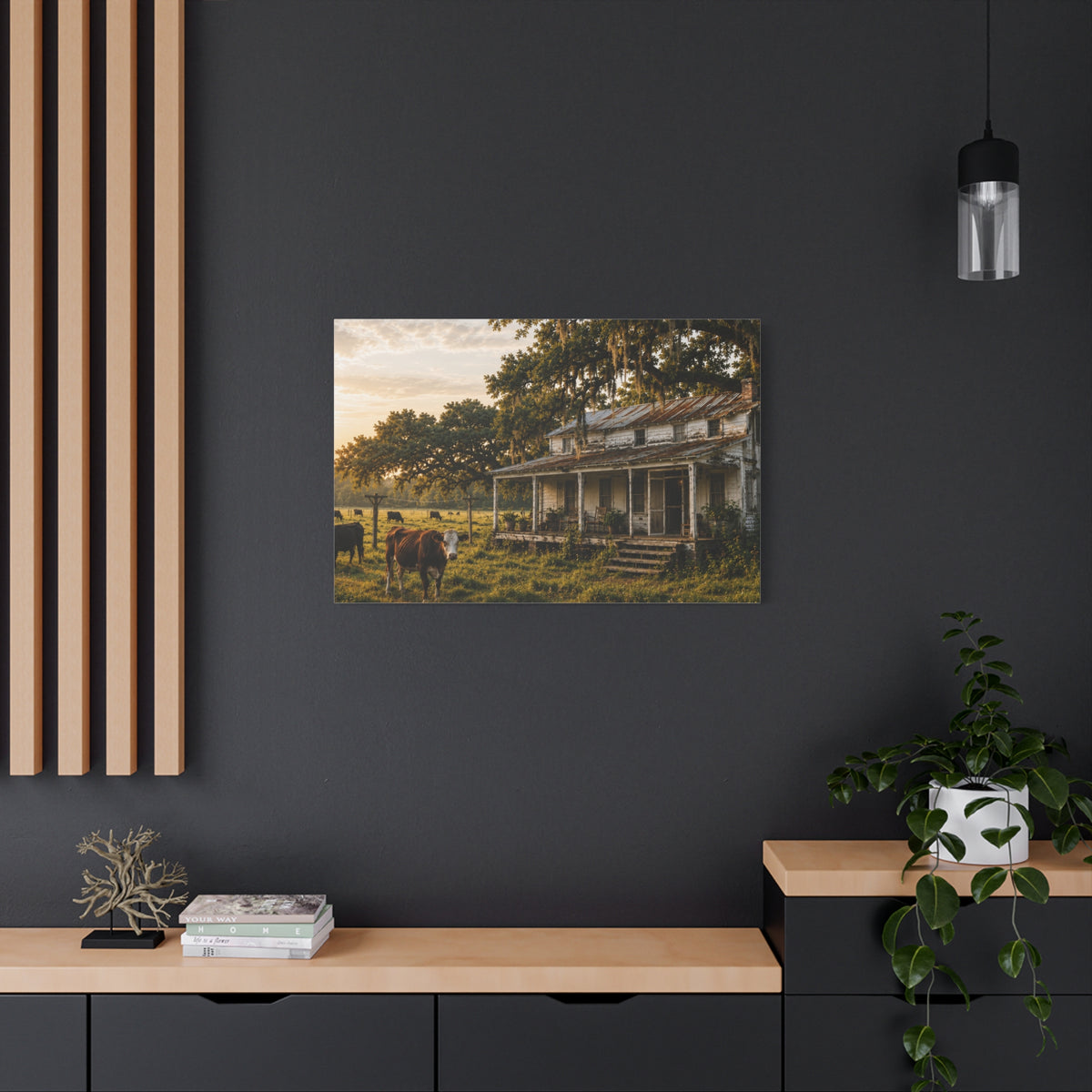 A rectangular The Good Old Homestead canvas print showing a weathered two-story farmhouse with a front porch in a grassy pasture, with a cow in the foreground, cattle, moss-draped oak trees, and warm sunset light, displayed on a dark charcoal wall above a light wood console with books, a plant, and decor.