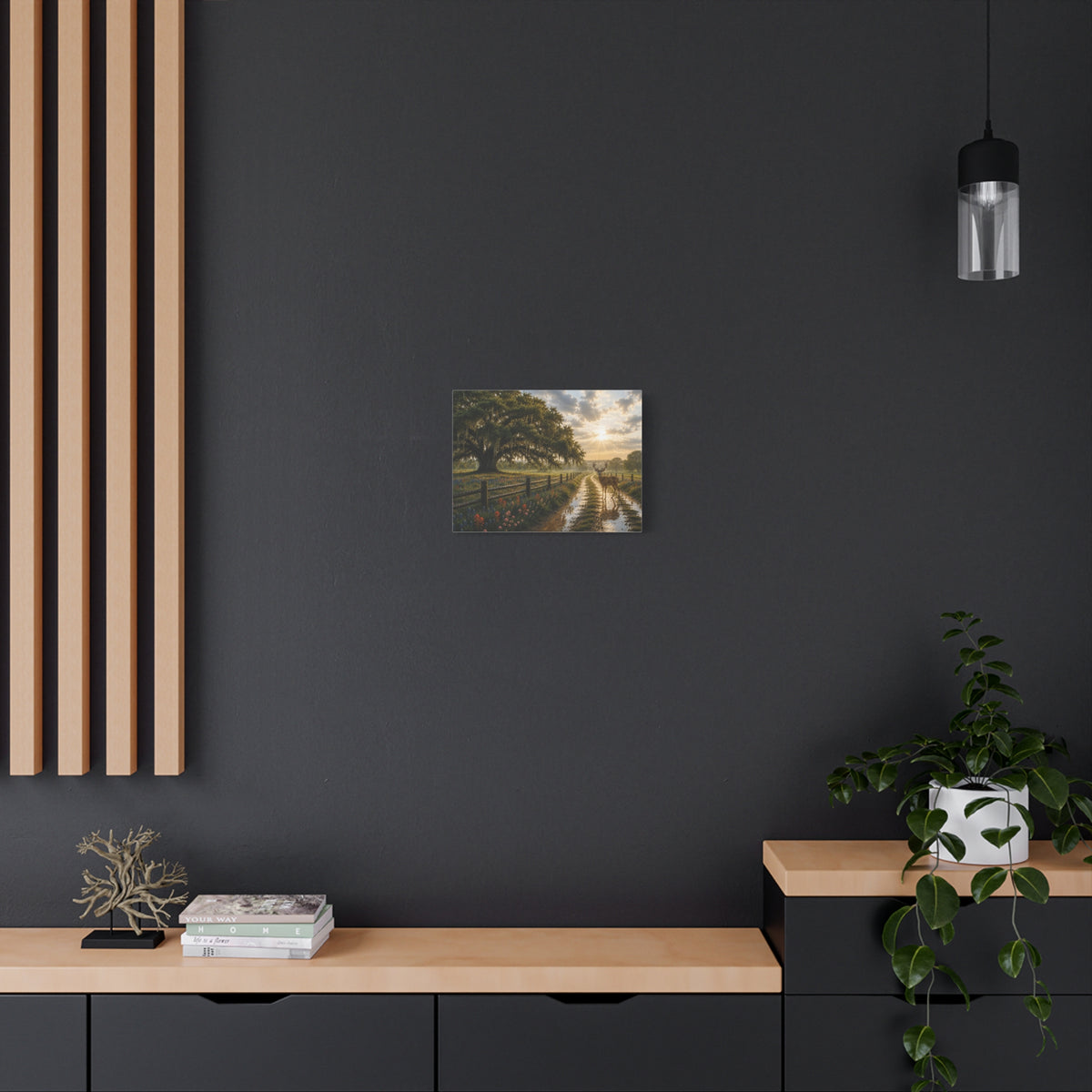 A small landscape art print depicting a tree-lined country road is displayed on a dark charcoal wall in a modern room setting with a wooden console, a potted plant, and a hanging light fixture.