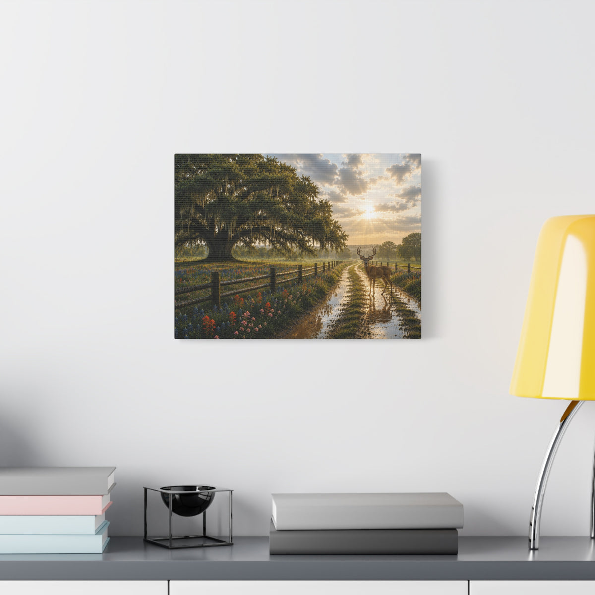A rectangular wall art print showing a sunlit country road with a large tree, fence, flowers, and a deer, displayed on a white wall above a shelf with books and a yellow lamp.