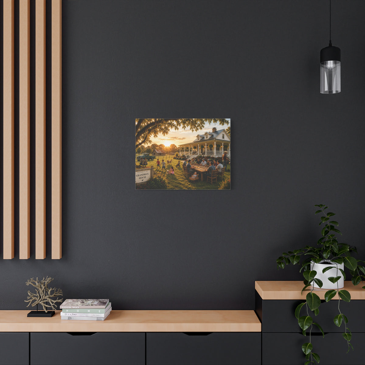A Grandma's House canvas print showing a family gathering outside a white farmhouse at sunset, displayed on a white wall above a console with a yellow lamp, books, and white flowers.