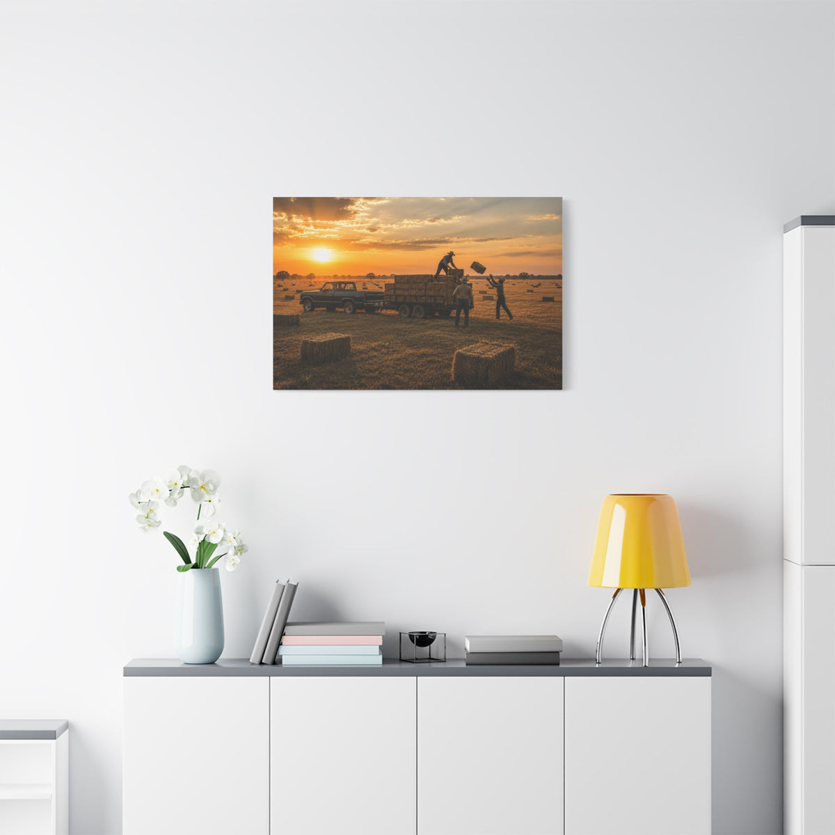 A Hay Season canvas print showing farmworkers loading rectangular hay bales onto a flatbed trailer beside a pickup truck in a golden field at sunset, displayed on a white wall above a console with a yellow lamp, books, and white flowers.