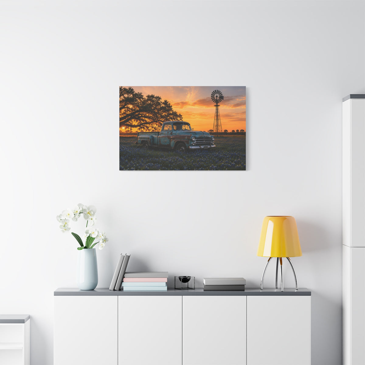 A square The Windmill canvas print showing a weathered blue vintage pickup in a field of bluebonnets at sunset, with a windmill and large oak tree in the background, displayed on a dark charcoal wall above a light wood console with books and a plant.