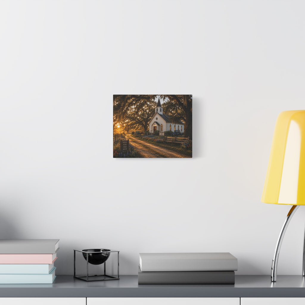A square The Country Church canvas print showing a small white country church with a steeple beside a dirt path, framed by moss-draped oak trees, rustic benches, wildflowers, and warm sunset light, displayed on a white wall above books and a yellow lamp.