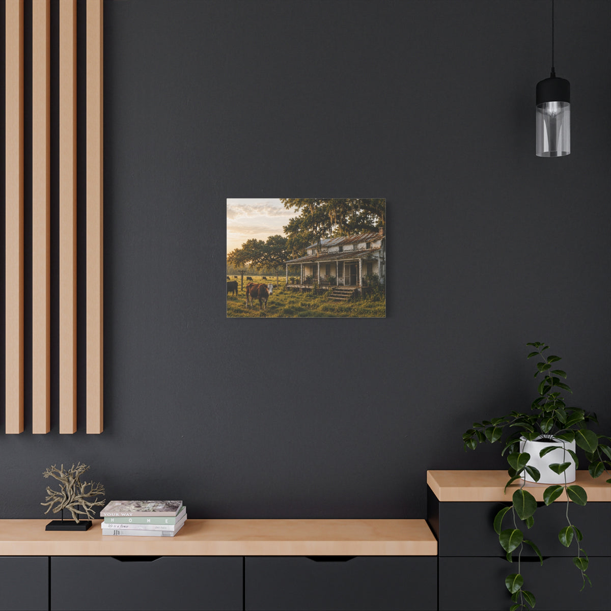 A square The Good Old Homestead canvas print showing a weathered two-story farmhouse with a front porch in a grassy pasture, with a cow in the foreground, cattle, moss-draped oak trees, and warm sunset light, displayed on a dark charcoal wall above a light wood console with books and a plant.