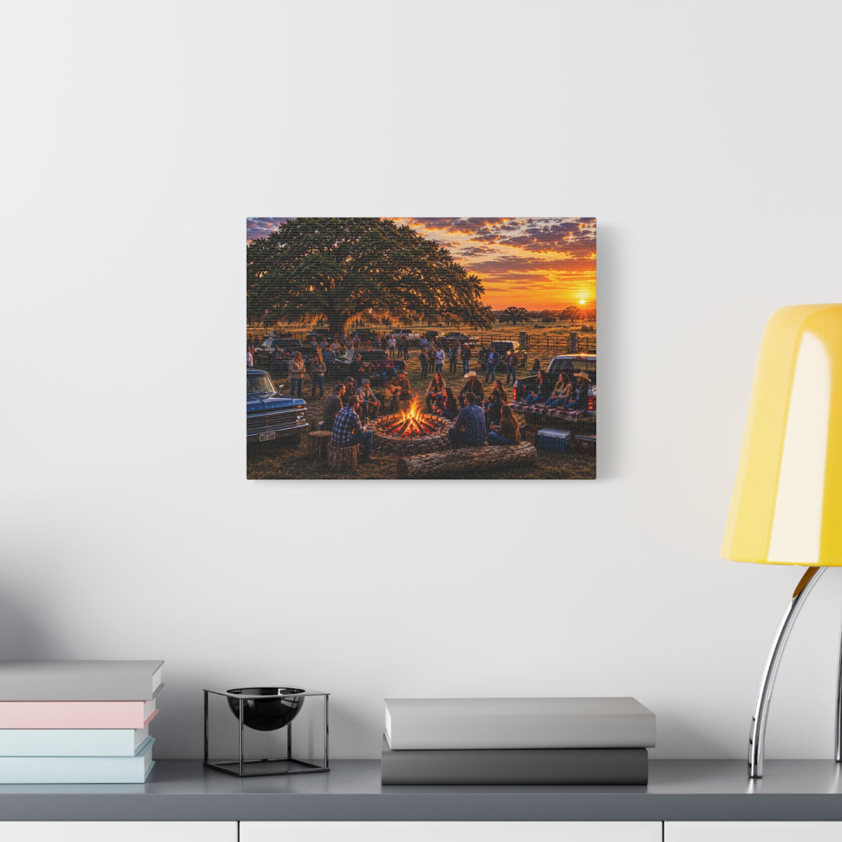 A Campfire Stories canvas print showing friends gathered around a ranch campfire beneath a large oak at sunset, displayed on a dark wall above a wooden shelf with a plant.
