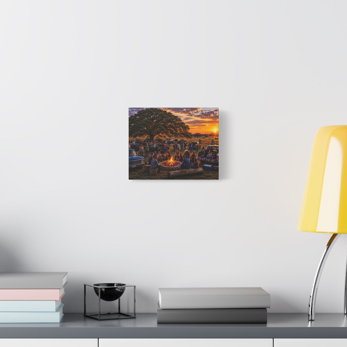 A square wall art print depicting a sunset campfire scene with people gathered under a tree, displayed on a white wall above a shelf with books and a yellow lamp.