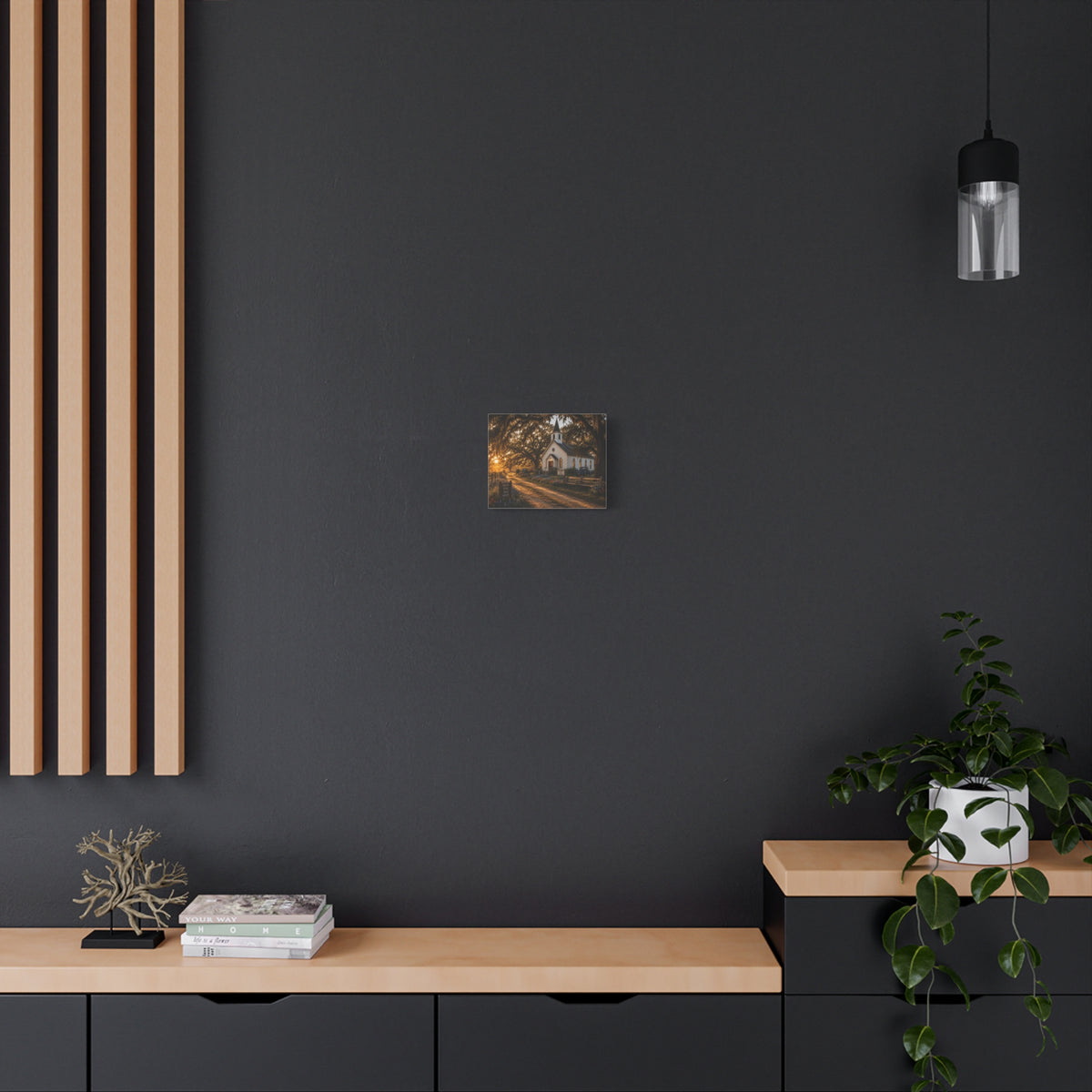 A The Country Church canvas print showing a small white country church with a steeple beside a dirt path, framed by moss-draped oak trees, rustic benches, wildflowers, and warm sunset light, displayed on a dark wall above wooden shelves with a plant and decor.