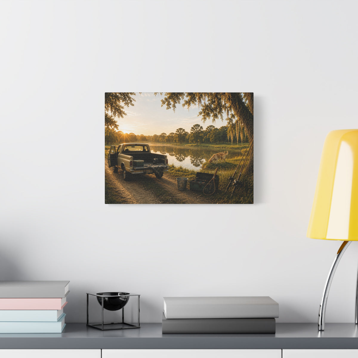 A rectangular The Old Fishing Hole canvas print showing a vintage pickup parked beside a quiet pond at sunset, with a dog, fishing rods, tackle, a cooler, and a moss-draped tree, displayed on a white wall above books, a black accent, and a yellow lamp.