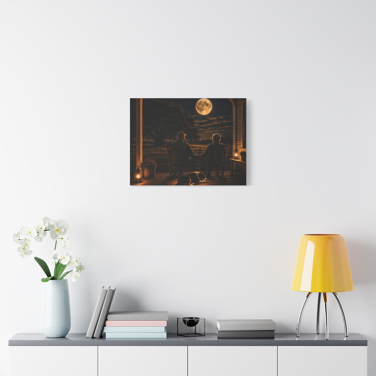A Harvest Moon canvas print showing an older couple holding hands in rocking chairs on a farmhouse porch with a sleeping dog, glowing lanterns, and a full moon over the fields, displayed on a white wall above a console with a yellow lamp, books, and white flowers.
