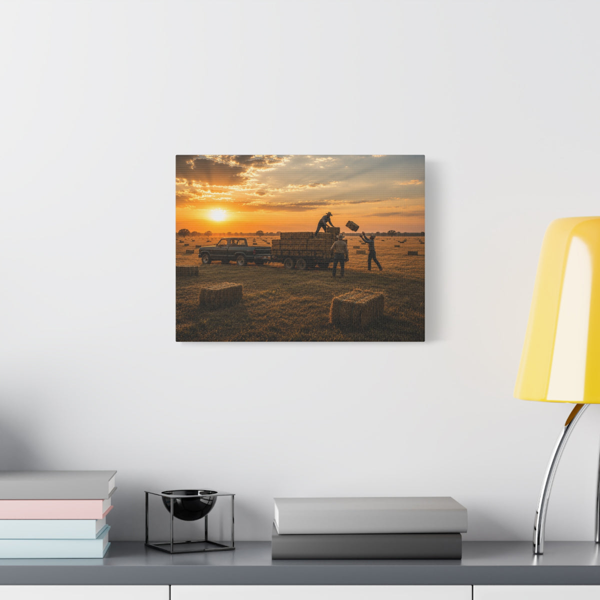 A rectangular Hay Season canvas print showing farmworkers loading rectangular hay bales onto a flatbed trailer beside a pickup truck in a golden field at sunset, displayed on a white wall above books, a black accent, and a yellow lamp.
